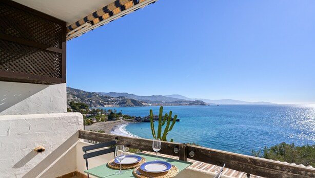 Un balcón con una mesa lista para dos personas, con vistas a la costa, montañas y un cactus.