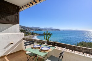 Un balcon avec une table dressée pour deux personnes, offrant une vue sur la côte, avec des montagnes et un cactus en arrière-plan.