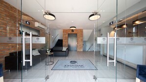 Un hall d’hôtel moderne avec des murs en verre, un mur d’accent en briques, un canapé gris et un escalier menant à l’étage supérieur.
