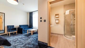 A modern hotel room with a living area featuring a sofa and chairs, a small table, and a glass-enclosed shower.