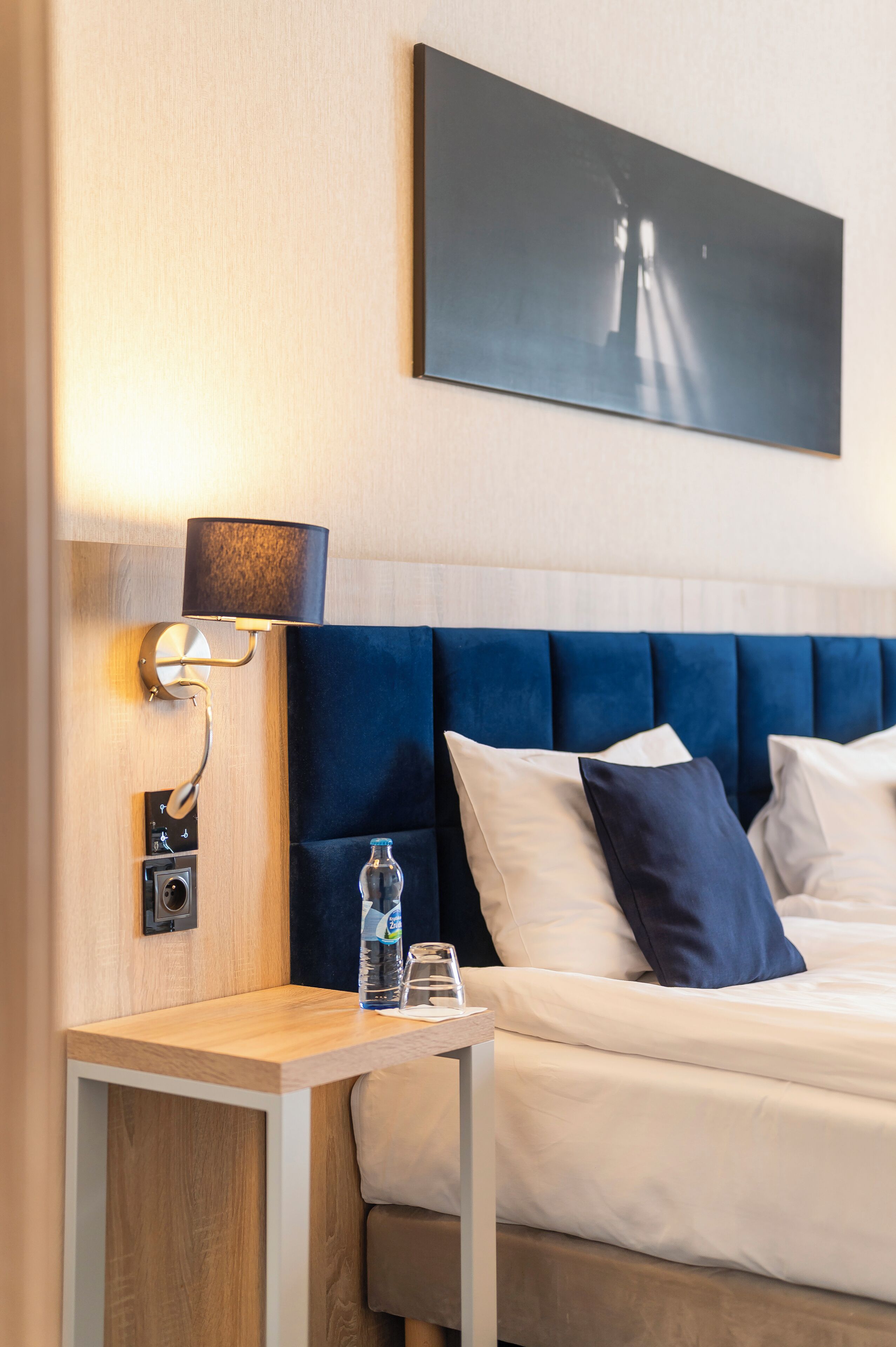 Une chambre d’hôtel avec un lit, une table de chevet avec une lampe, une bouteille d’eau et un tableau au mur.