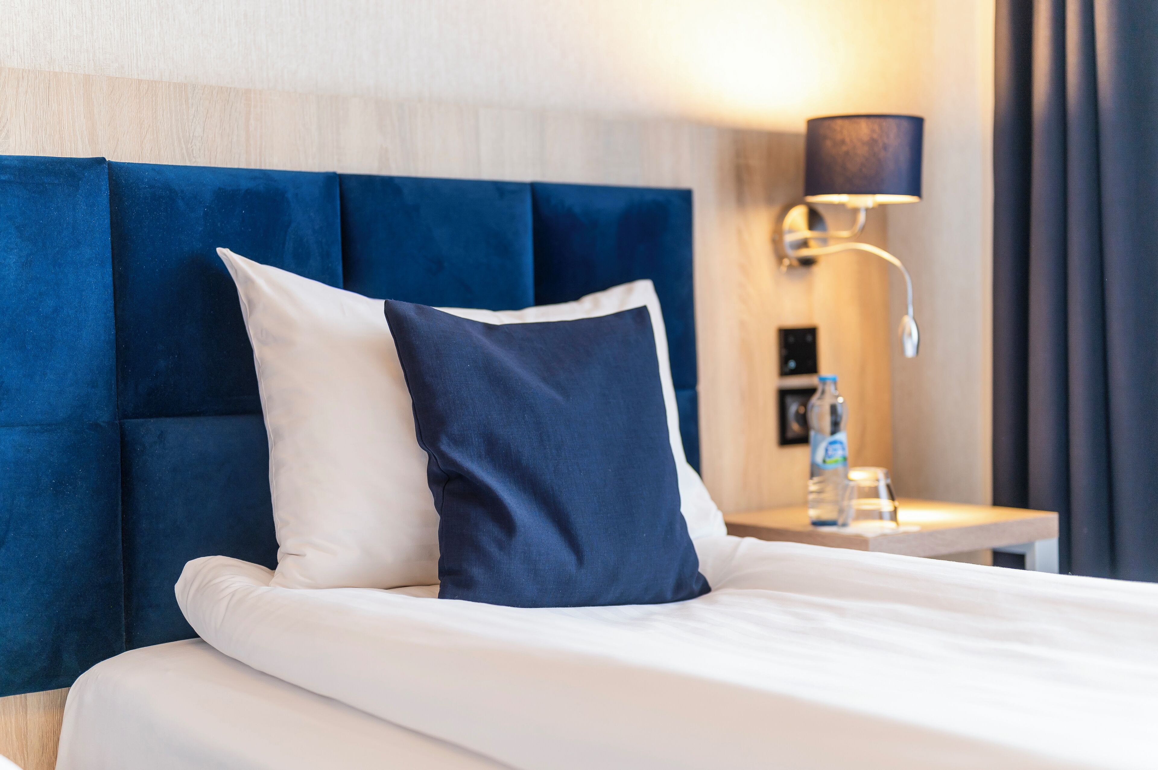 A hotel room with a bed, a blue upholstered headboard, a white pillow, a dark blue pillow, a bedside table with a lamp, and a bottle of water.