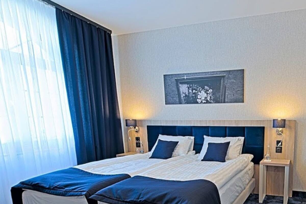 Een tweepersoonsbed met een donker hoofdbord, twee nachtkastjes met lampen, een groot raam met gordijnen en een ingelijst beeld aan de muur.
