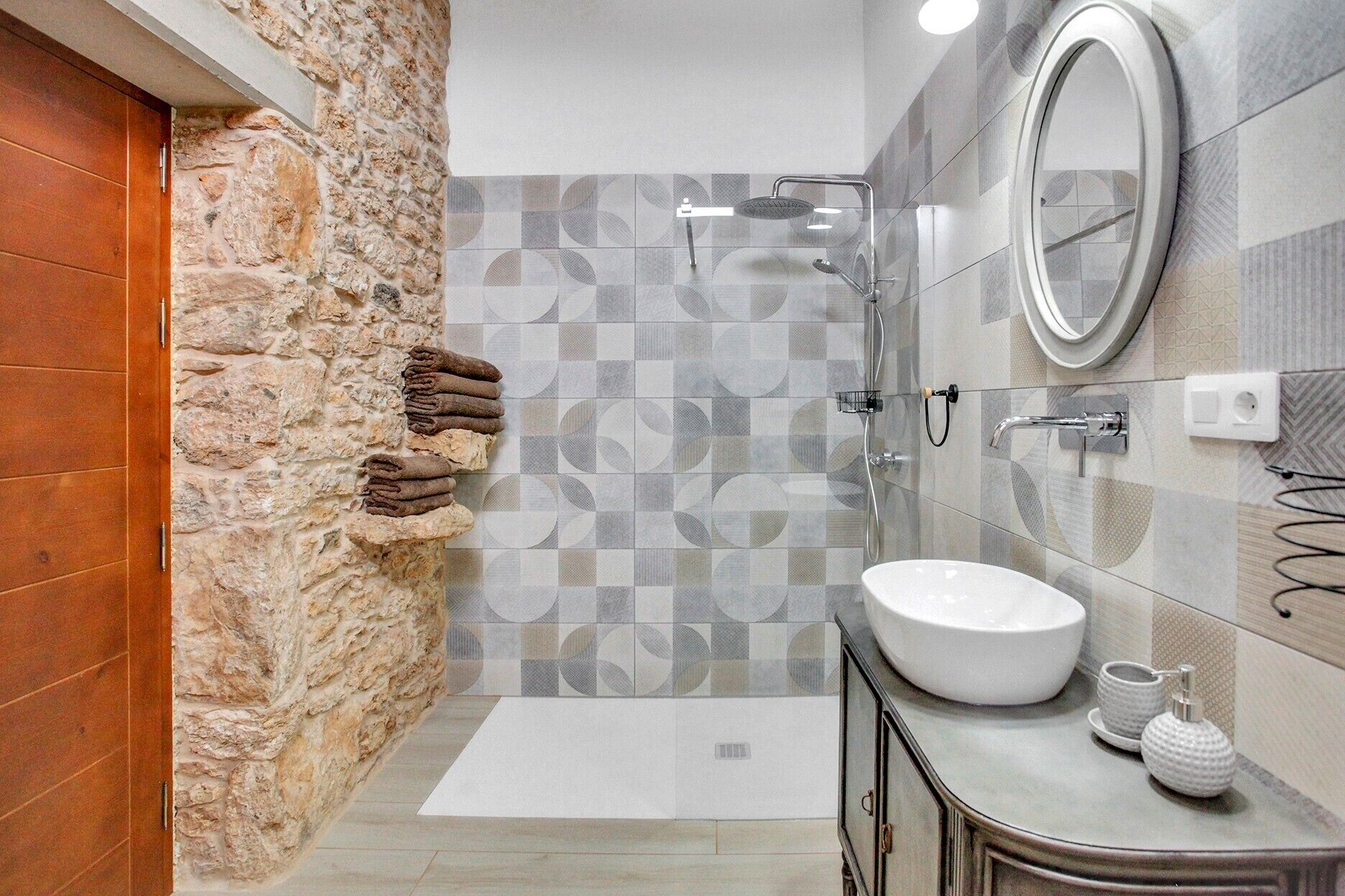 Un baño con pared de piedra, zona de ducha con azulejos, lavamanos y espejo.