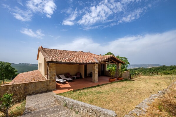 A stone house with a tiled roof, a covered patio with seating, and a view of the countryside.