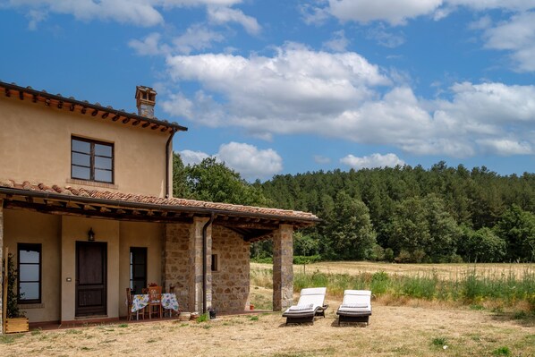 A two-story stone house with a tiled roof, a porch with a table and chairs, and two lounge chairs in the foreground.