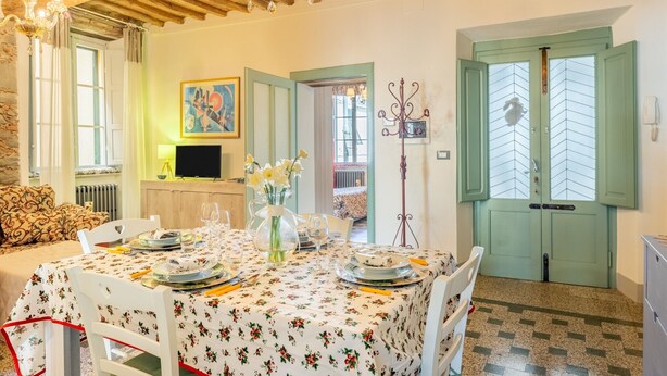 Un coin repas avec une table dressée pour quatre personnes, un téléviseur, un vase rempli de fleurs et une porte avec des volets verts.