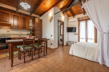 A kitchen with wooden cabinets, a dining table with chairs, a washing machine, and a bed with curtains.. Holiday Home "Dependance Dei Ginepri" with Sea View, Terrace & Wi-Fi