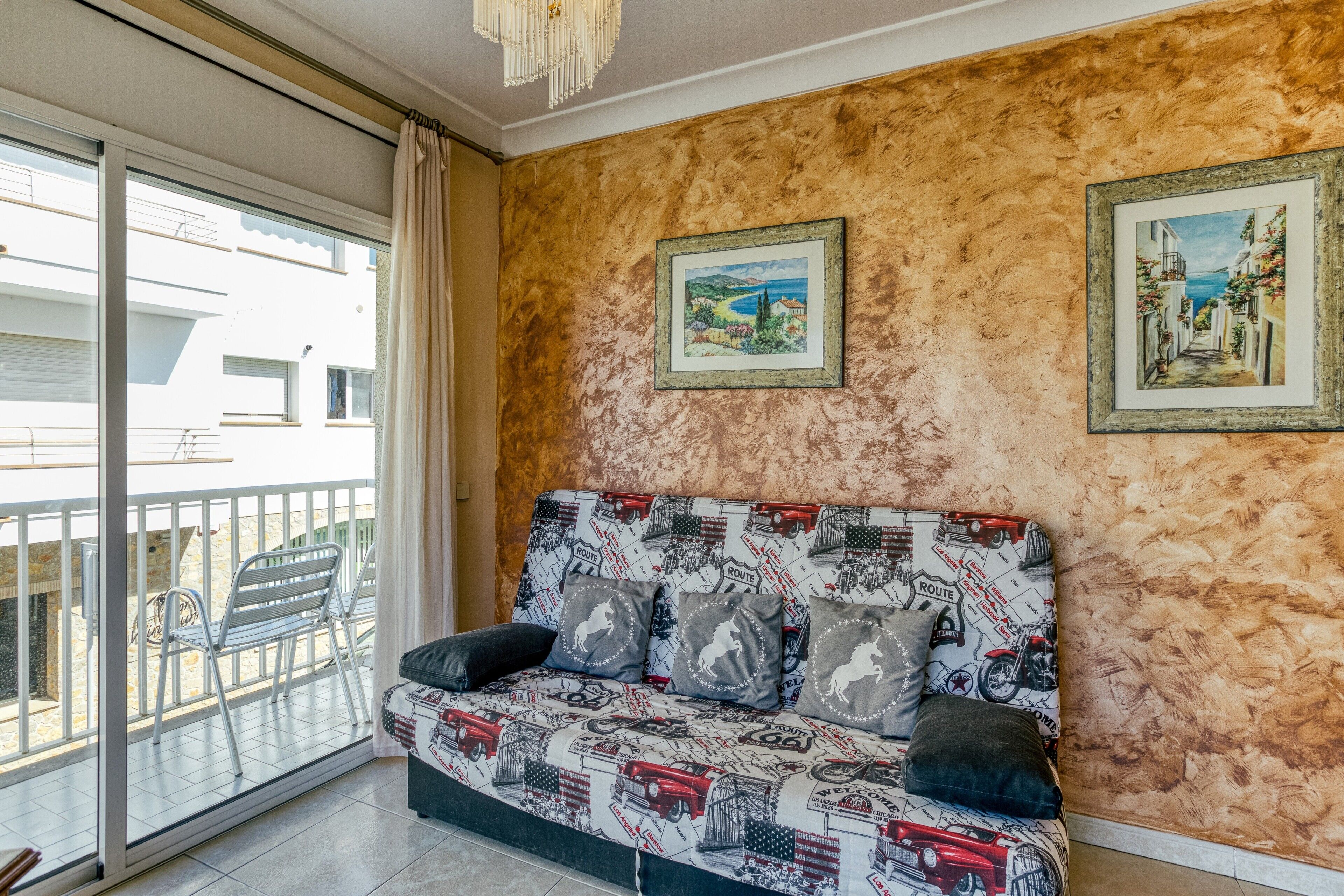 Une pièce avec un canapé, deux tableaux encadrés au mur, un balcon avec des chaises et une vue sur les bâtiments environnants.