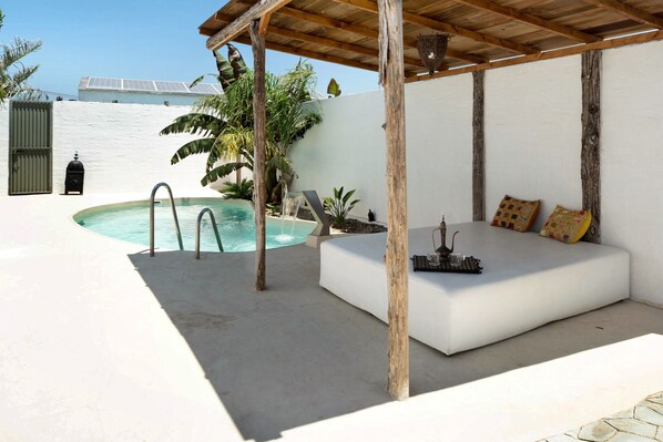 A pool area with a white outdoor seating area, a pergola, and a white wall with a gate.