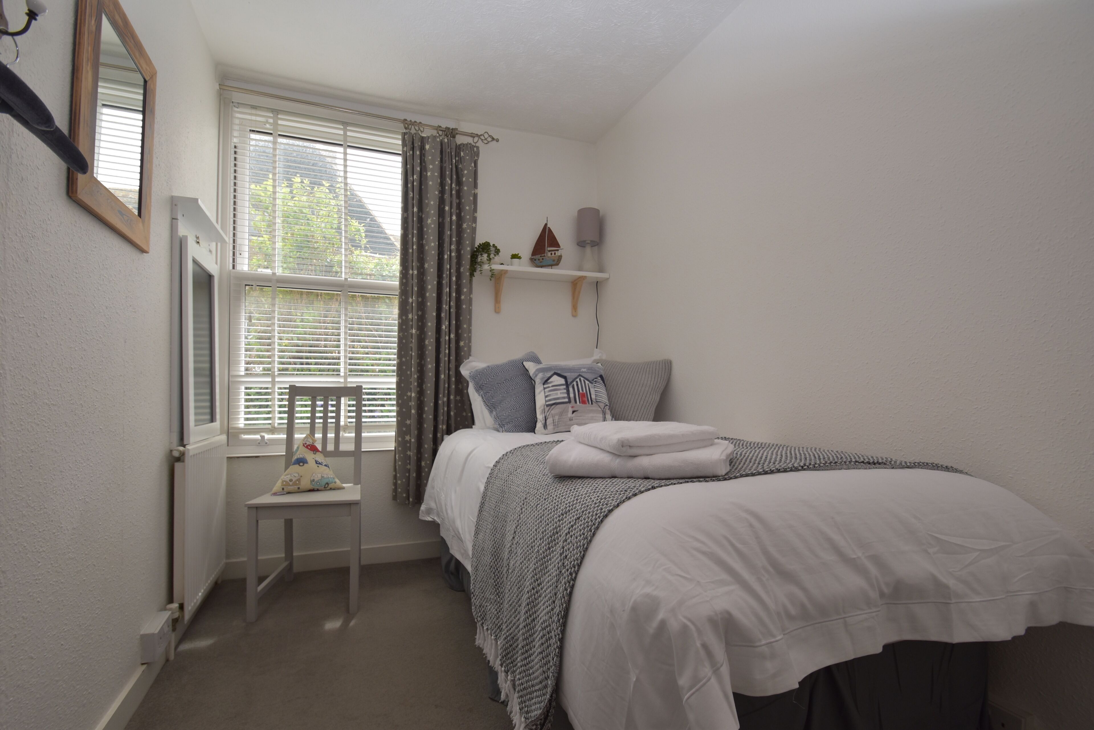 A bedroom with a bed, a chair, a window with curtains, a shelf with decorative items, and a mirror on the wall.