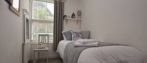 A bedroom with a bed, a chair, a window with curtains, a shelf with decorative items, and a mirror on the wall.