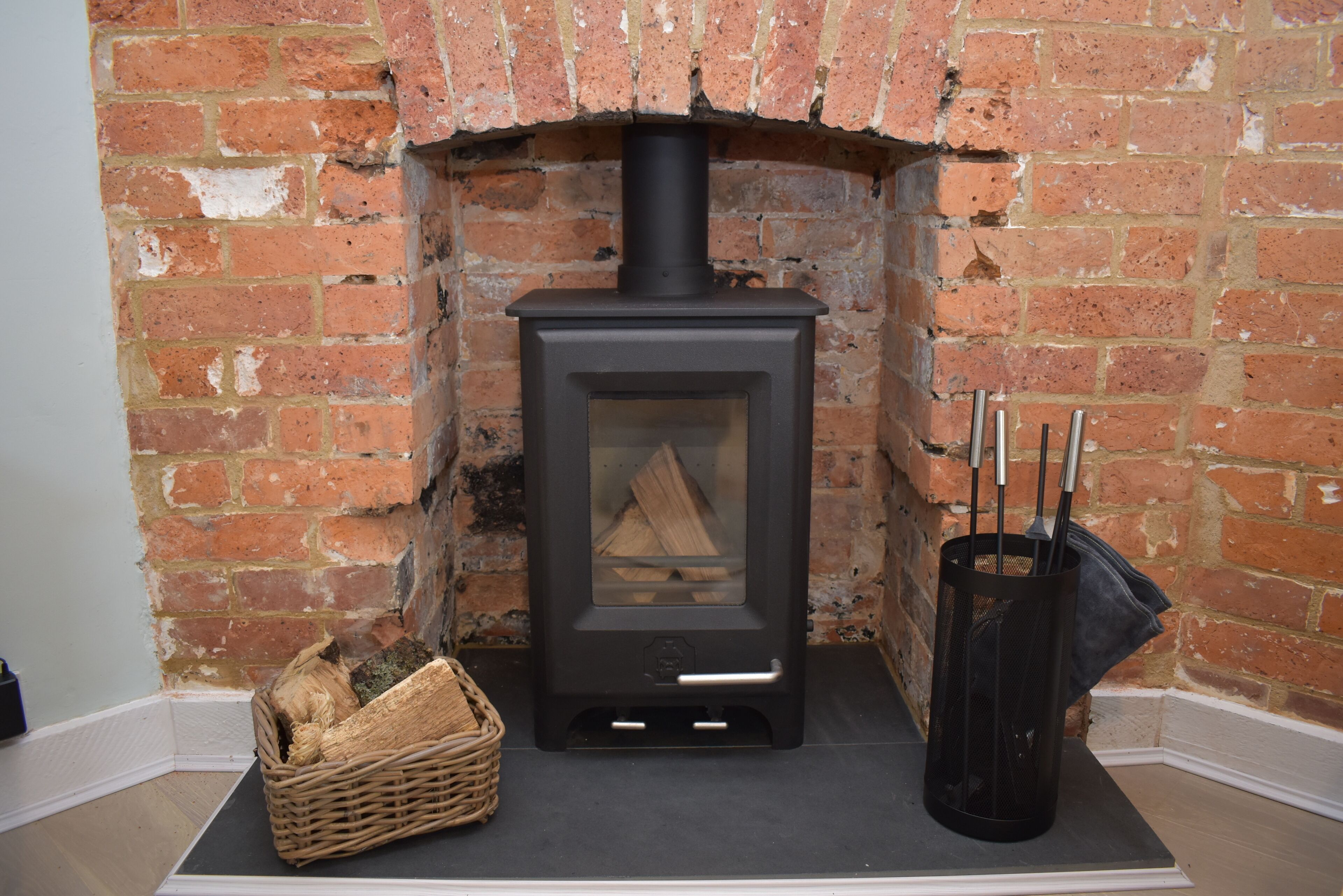 A wood-burning stove in a brick fireplace, a basket of firewood, and a metal ash collector.