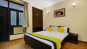 A bedroom with a bed, a nightstand, a wall-mounted lamp, a window with curtains, and a painting on the wall. - Itsy Hotels Arien International (New Delhi)