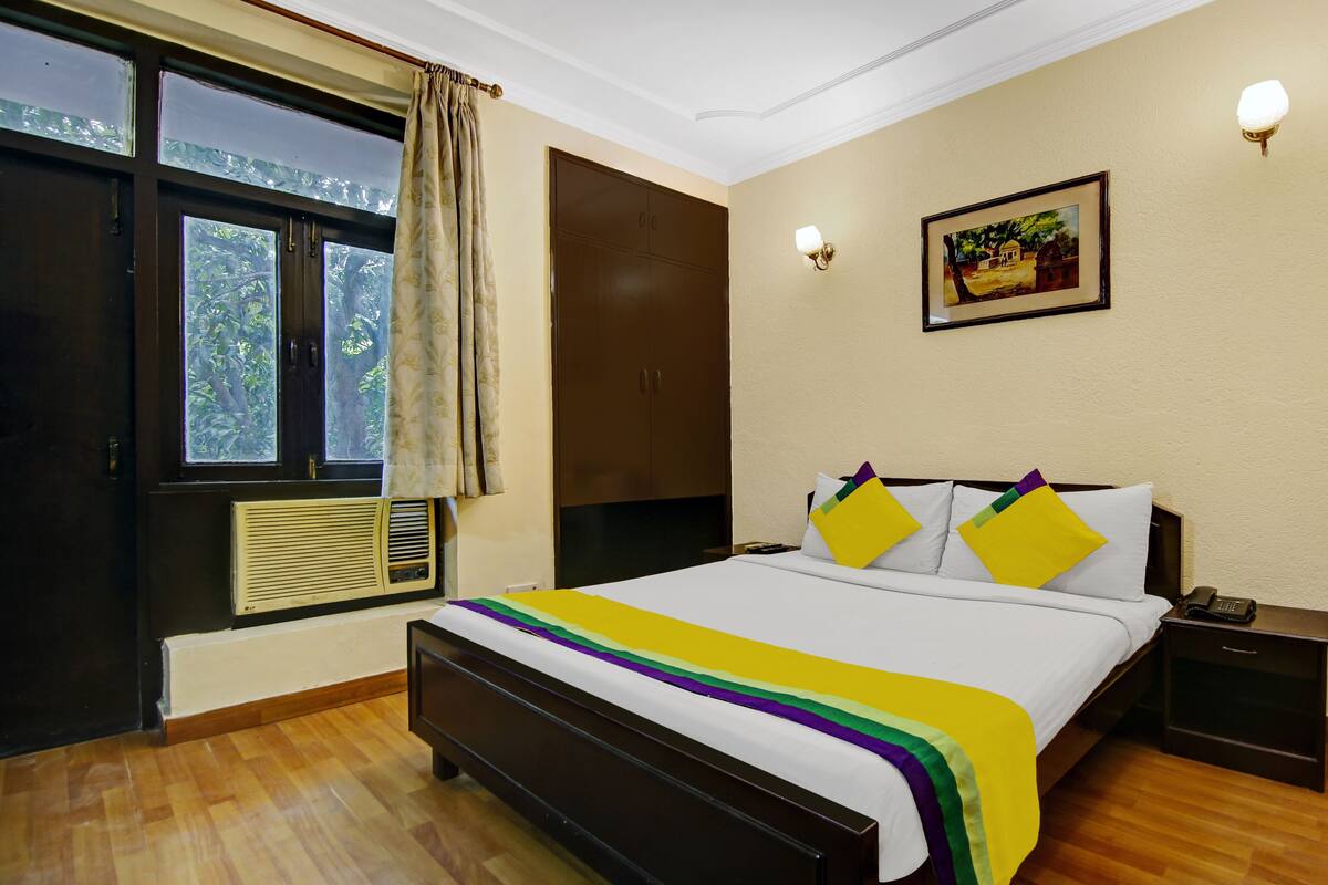 A bedroom with a bed, a nightstand, a wall-mounted lamp, a window with curtains, and a painting on the wall.