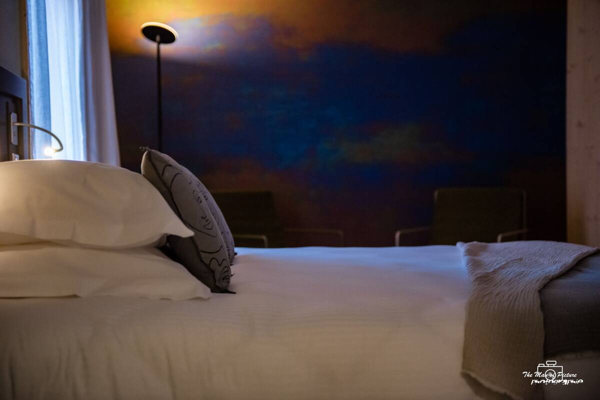 Una habitación de hotel con cama, almohadas, una lámpara de noche y un mural en la pared.