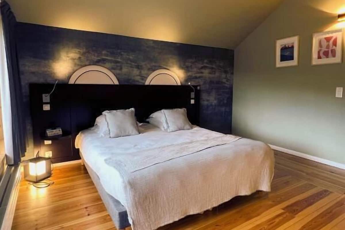 Un dormitorio con una cama grande, suelo de madera, cabecero oscuro e iluminación instalada en la pared.