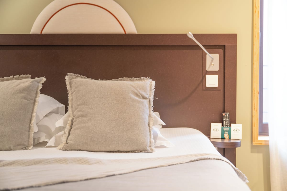 Una cama bien hecha con dos almohadas, una mesita de noche con un control remoto y un reloj de pared.