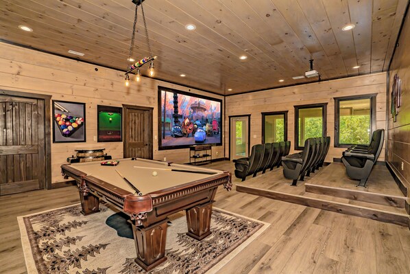 A pool table with a billiard cue, a television mounted on the wall, and a row of black chairs.