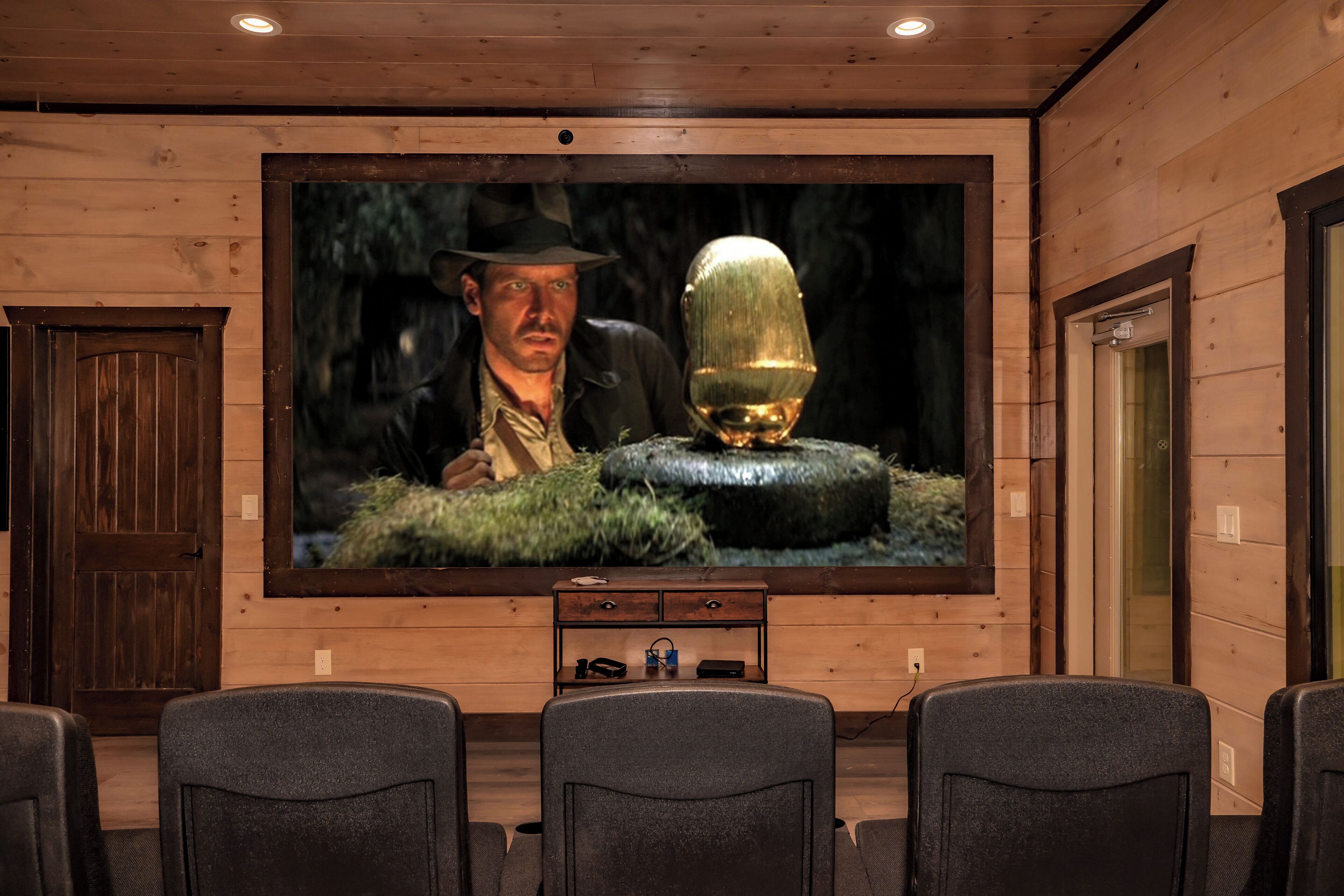 A wooden-paneled room with a large flat-screen TV mounted on the wall, a wooden door, and a small table with electronic devices.