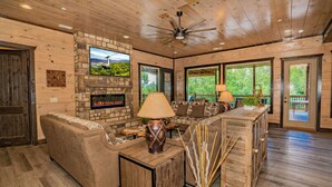 A spacious living room with a stone fireplace, wooden ceiling, and large windows. - Dreamers Mountain Retreat: Pool, Theater, Hot Tub, WiFi, Games, Fire Pit (Sevierville)