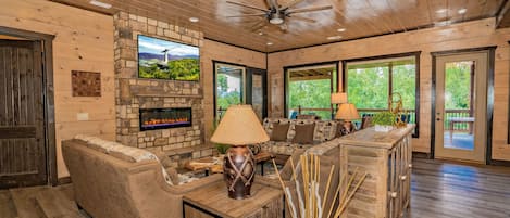 A spacious living room with a stone fireplace, wooden ceiling, and large windows.