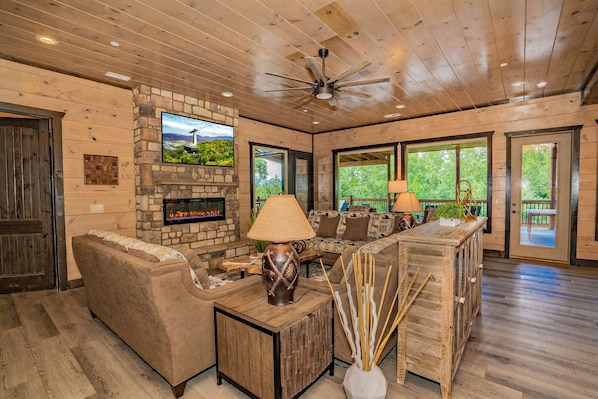 A spacious living room with a stone fireplace, wooden ceiling, and large windows.