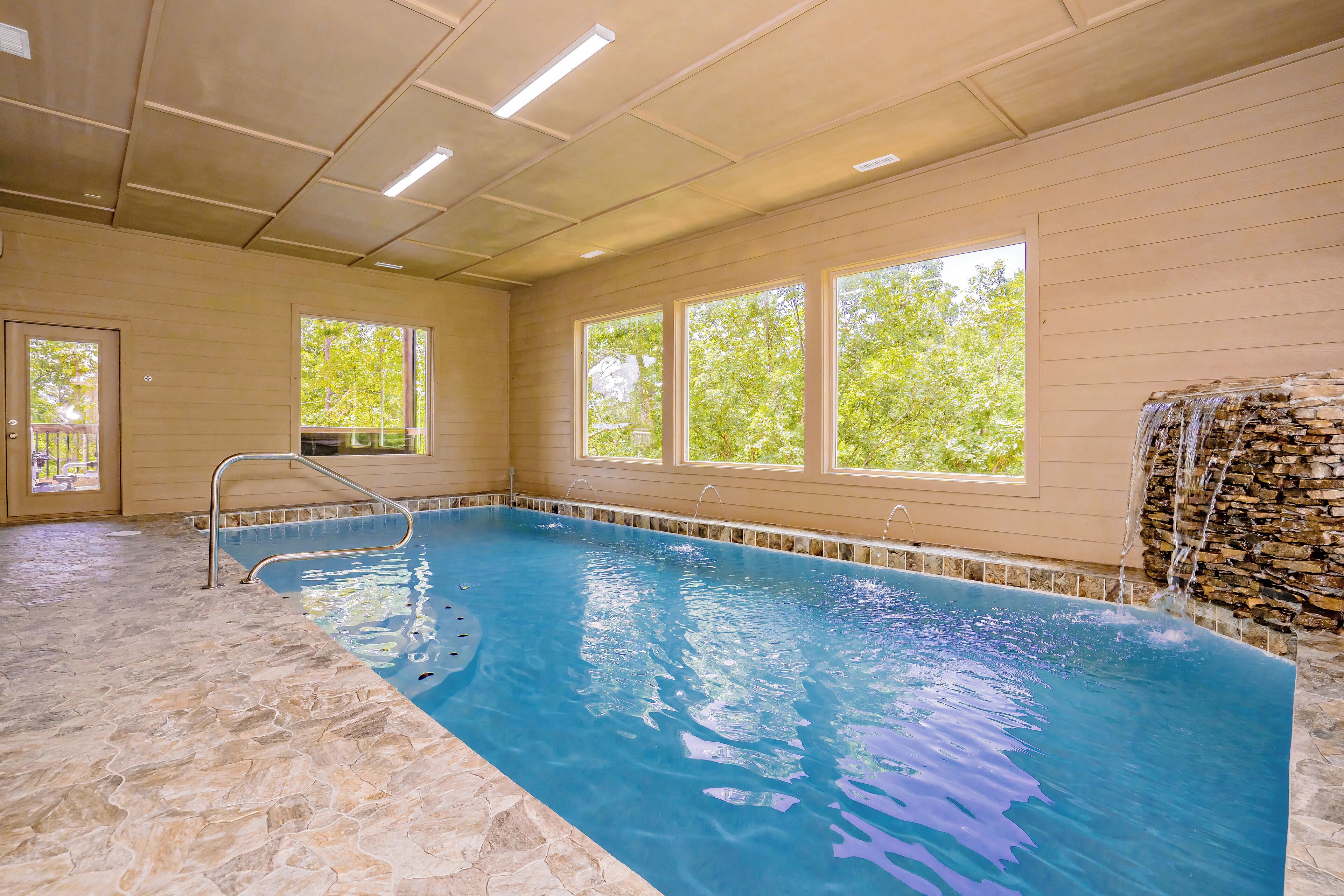 Una piscina interior con ventanas transparentes, suelo de piedra y una cascada de piedra.
