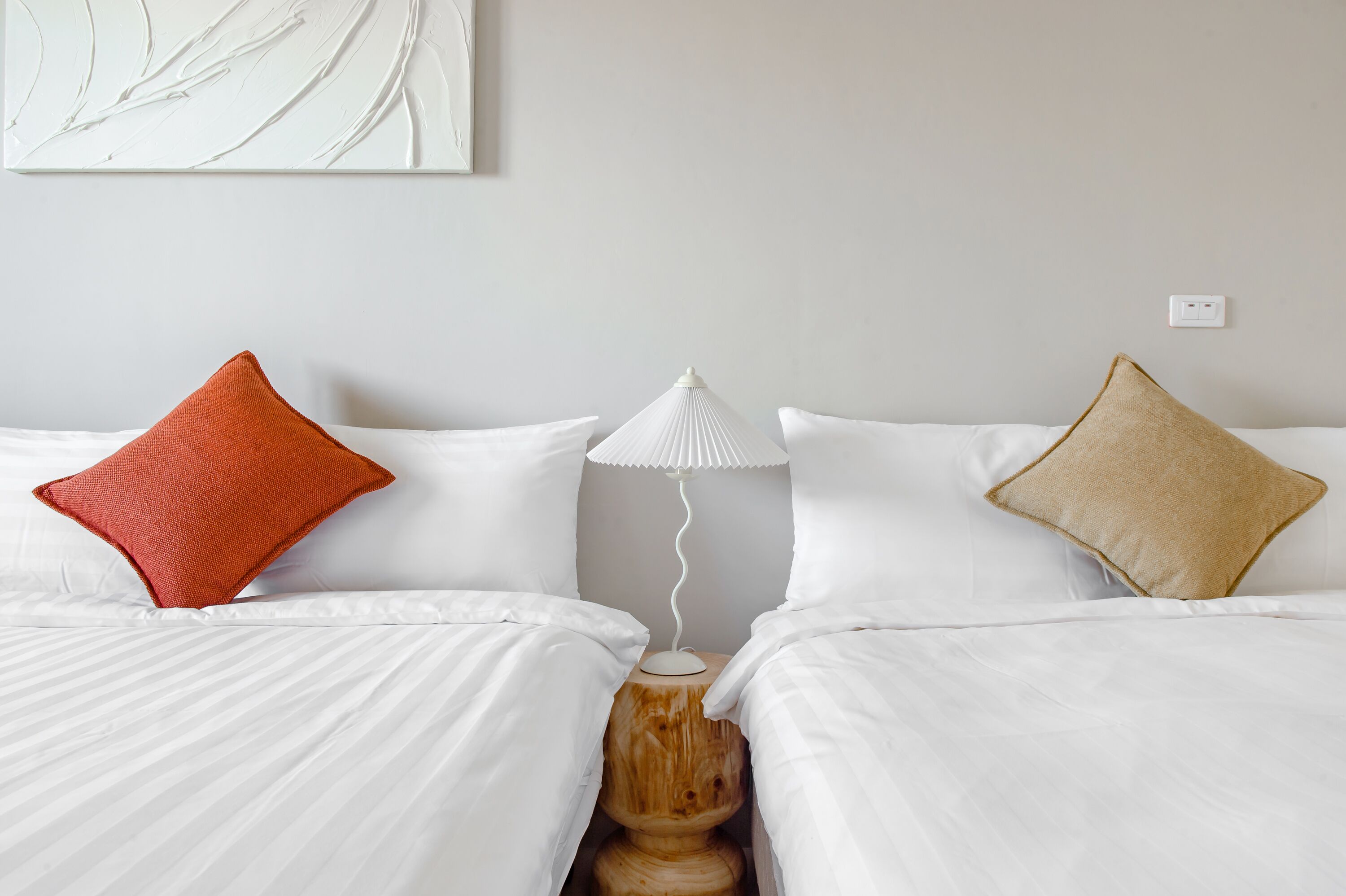 Habitación de hotel con dos camas, ropa de cama blanca, una mesita de noche de madera y una lámpara blanca entre las camas.