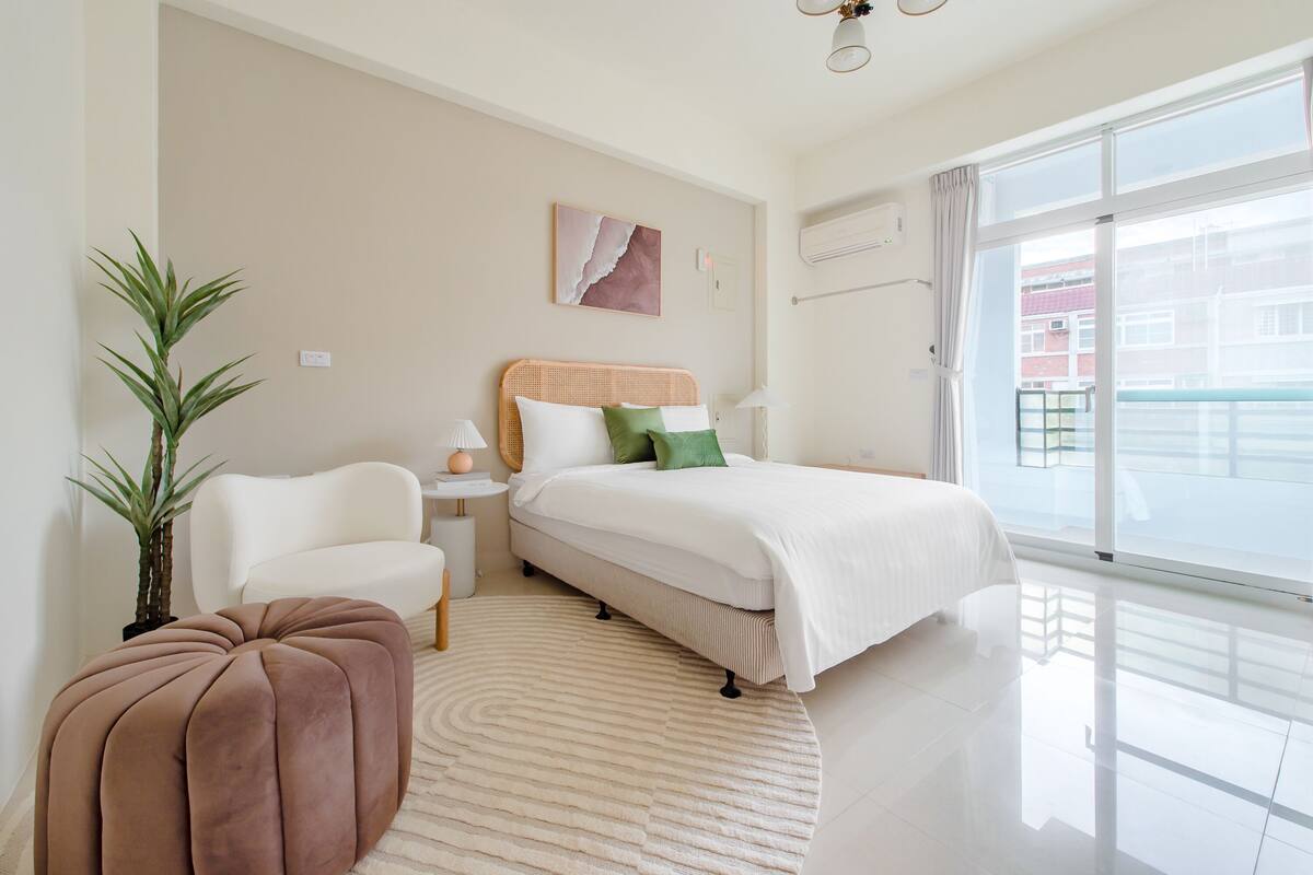 Una habitación de hotel moderna con una cama, un sillón blanco, un taburete redondo, una mesita con lámpara, aire acondicionado de pared y un ventanal con vistas a edificios.
