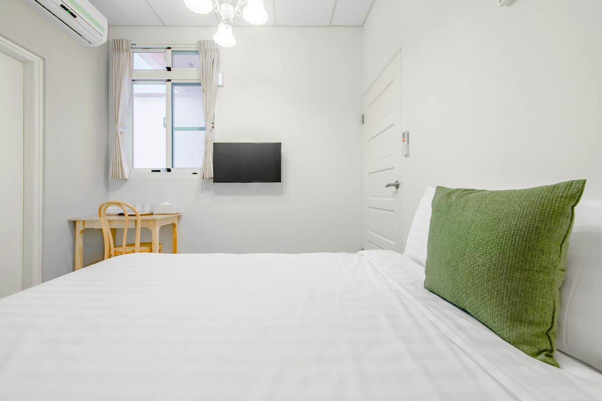 Un dormitorio con cama, escritorio, silla, televisor y ventana con cortinas.