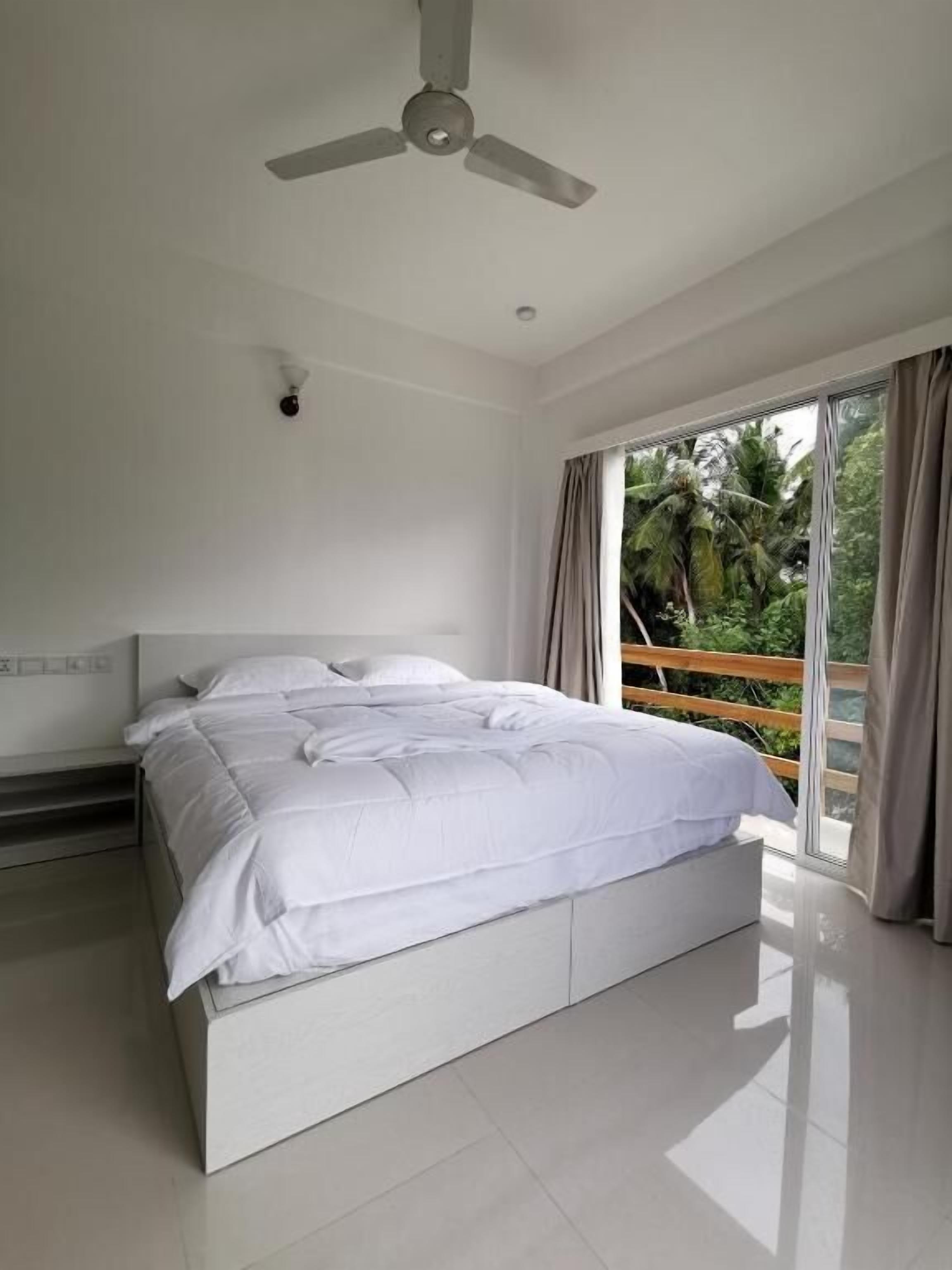 A modern bedroom with a bed, a ceiling fan, and a view of greenery through large windows.