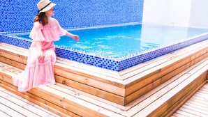 Una persona con vestido rosa sentada en escalones de madera junto a una piscina con mosaicos azules.