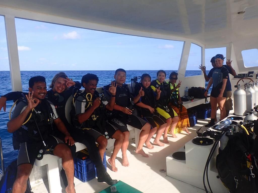 Un grupo de personas en un bote con equipo de buceo, sentadas en un banco y una mesa, con el océano al fondo.