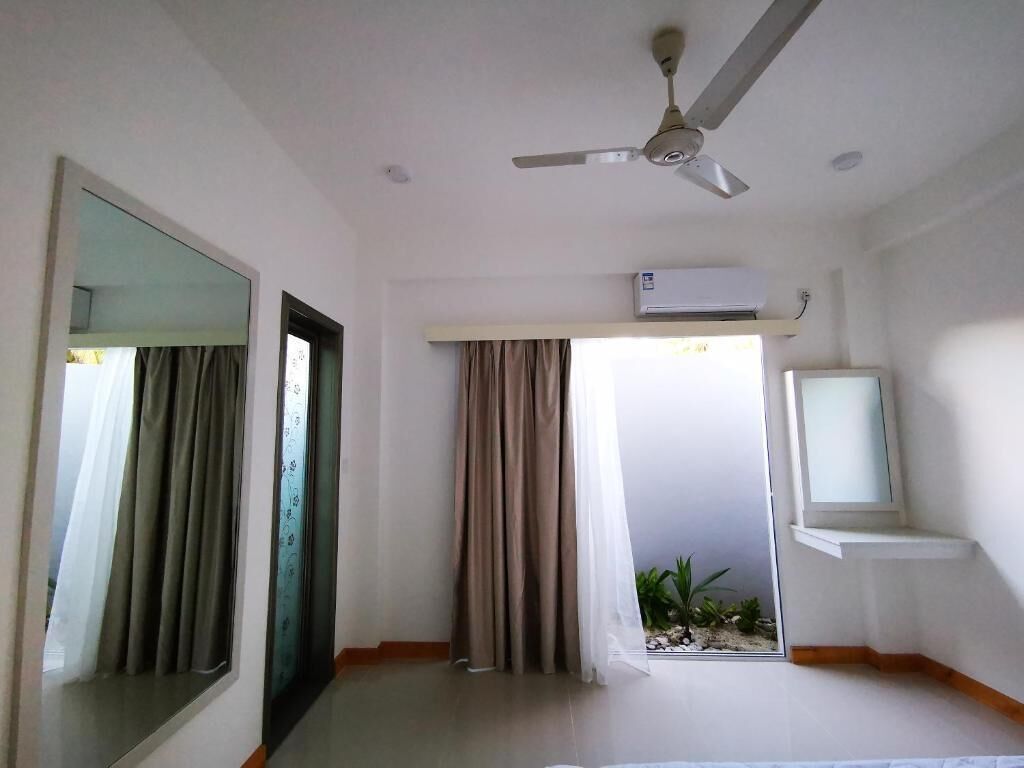 A hotel room with a ceiling fan, an air conditioning unit, a television on a stand, and a window with curtains.