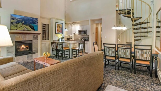 A spacious living room with a fireplace, a dining area, and a kitchen in the background. A spiral staircase is visible to the right. - Aspen at Streamside at Vail (Vail)