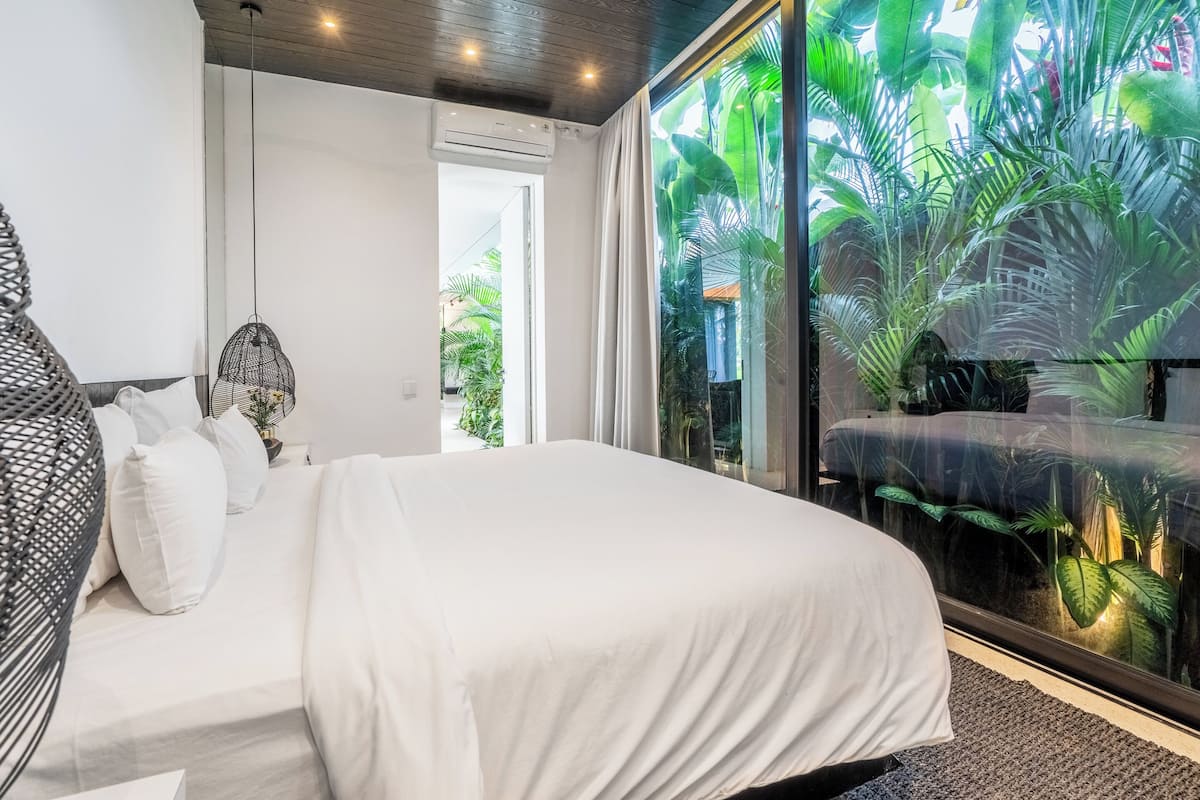 Un dormitorio moderno con una cama grande, una silla colgante y vistas a un follaje exuberante a través de un muro de cristal.