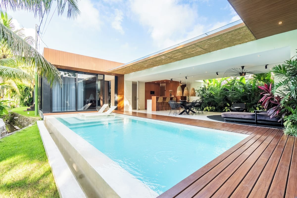Un área de piscina moderna al aire libre con cubierta de madera, un salón con paredes de vidrio y exuberante vegetación tropical.