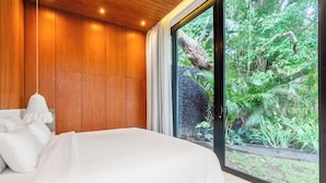 A bedroom with a large bed, wooden panel walls, a hanging light fixture, and a view of a lush garden through a sliding glass door. - Green Flow Villa 14 (Ubud)