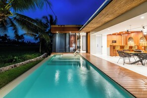 Un área moderna junto a la piscina con terraza de madera, piscina y zona de comedor con sillas y mesa.