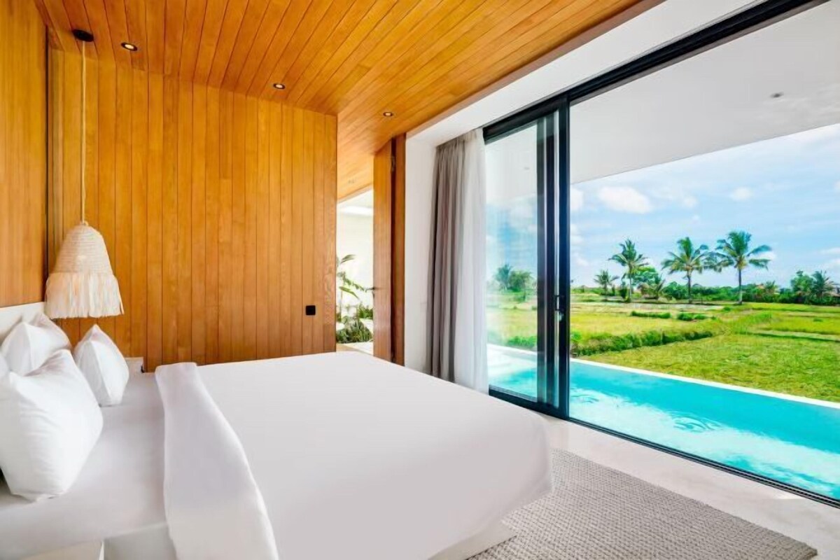 Une chambre avec un grand lit, des murs recouverts de panneaux de bois, un luminaire suspendu et une vue sur une piscine et un espace verdoyant.