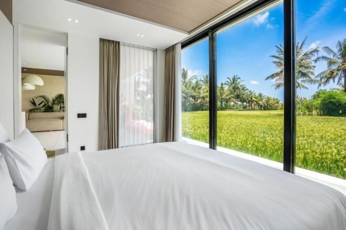 Une chambre avec un grand lit, une fenêtre donnant sur un champ verdoyant et des palmiers, et une salle de bain équipée d’un porte-serviettes.