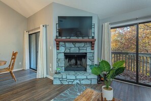 A living room with a stone fireplace, a television mounted above it, a dining area with a table and chairs, and a large window with a view of trees.