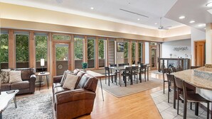 A spacious living area with a large dining table, comfortable seating, and large windows offering views of the outdoors.