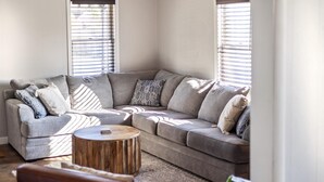Una sala de estar con rincón, un sofá seccional gris, cojines estampados, una mesa de centro de madera y ventanas amplias con persianas.