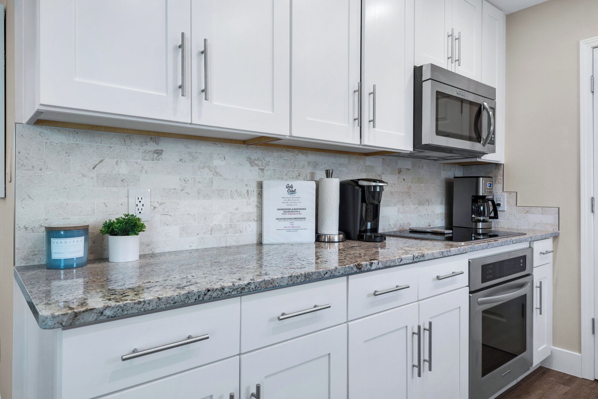 Una cocina moderna con armarios blancos, microondas de acero inoxidable, cafetera y horno empotrado.