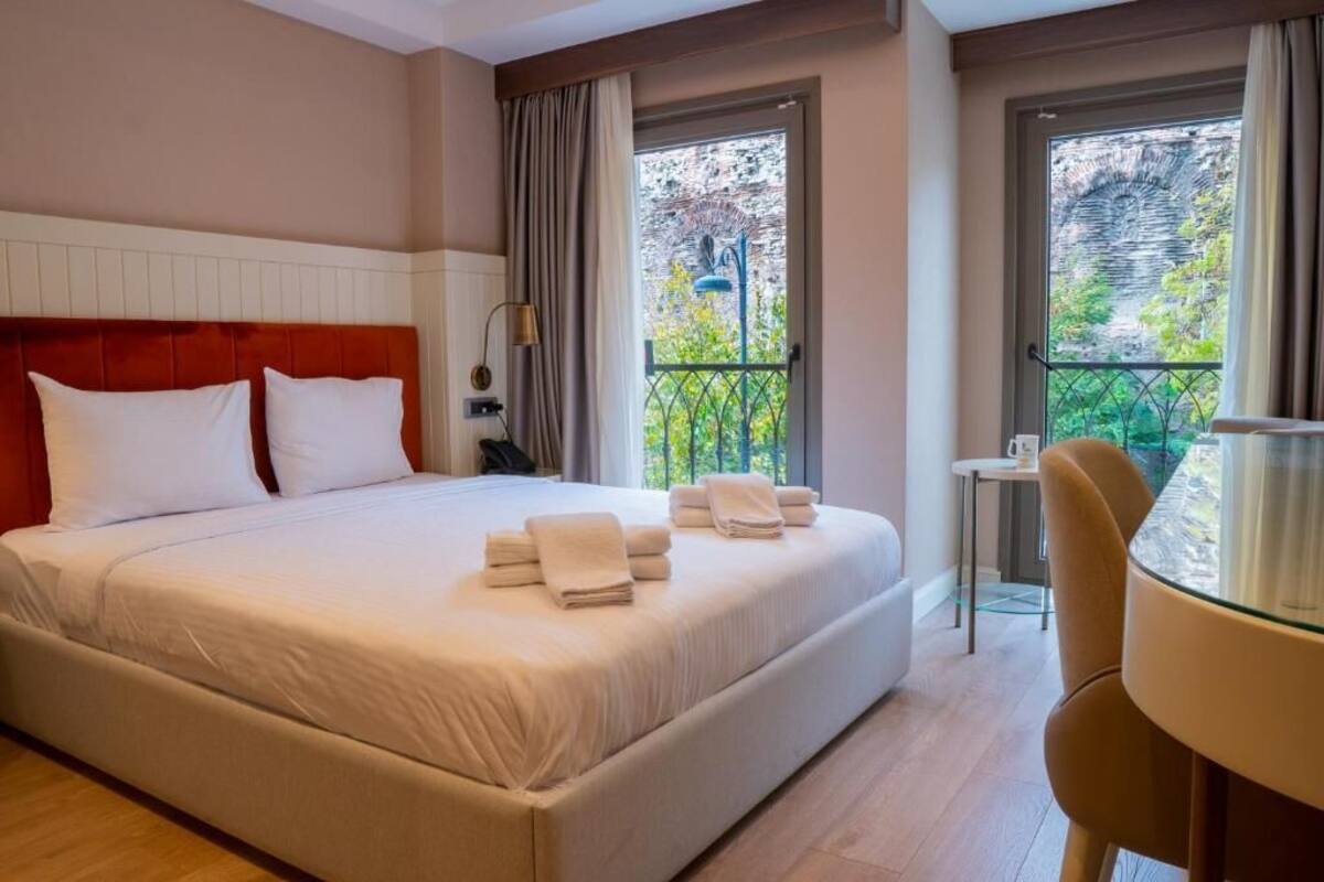 A hotel room with a large bed, a small table, and a chair. There are windows with a view of greenery.