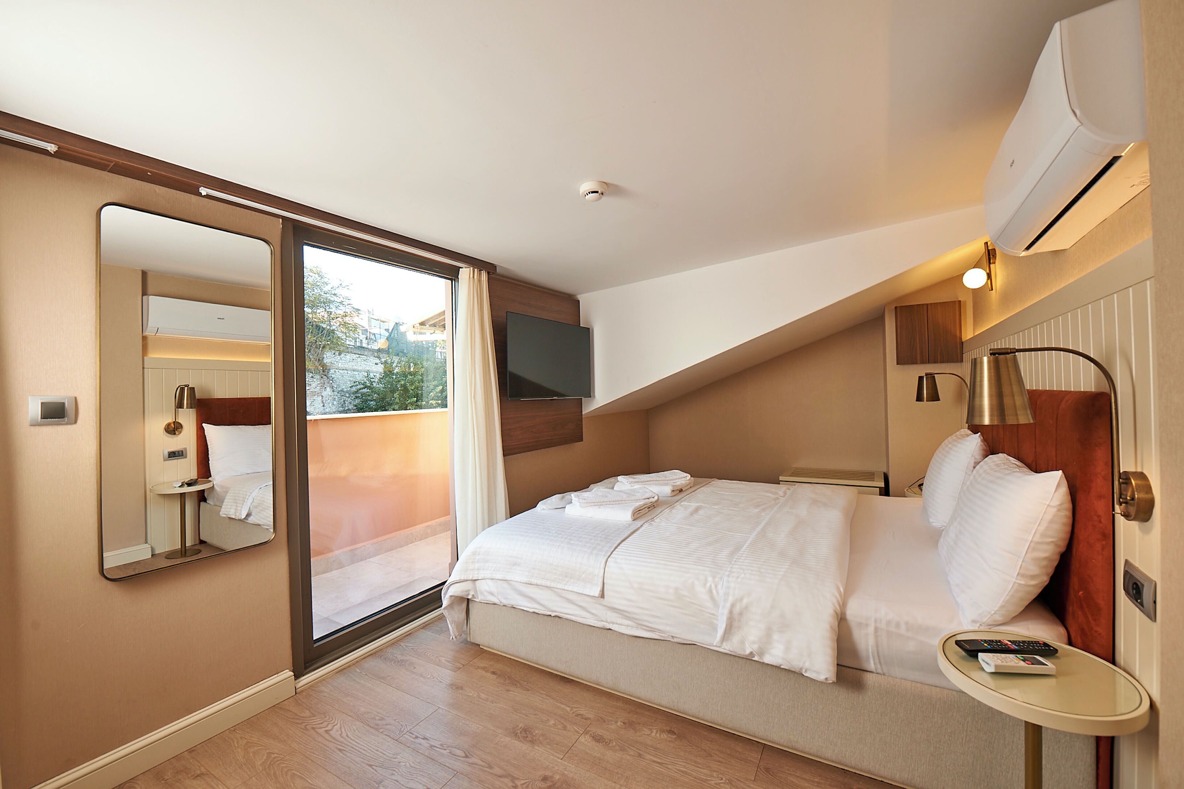 A hotel room with a bed, a small table, a mirror, and an air conditioning unit.