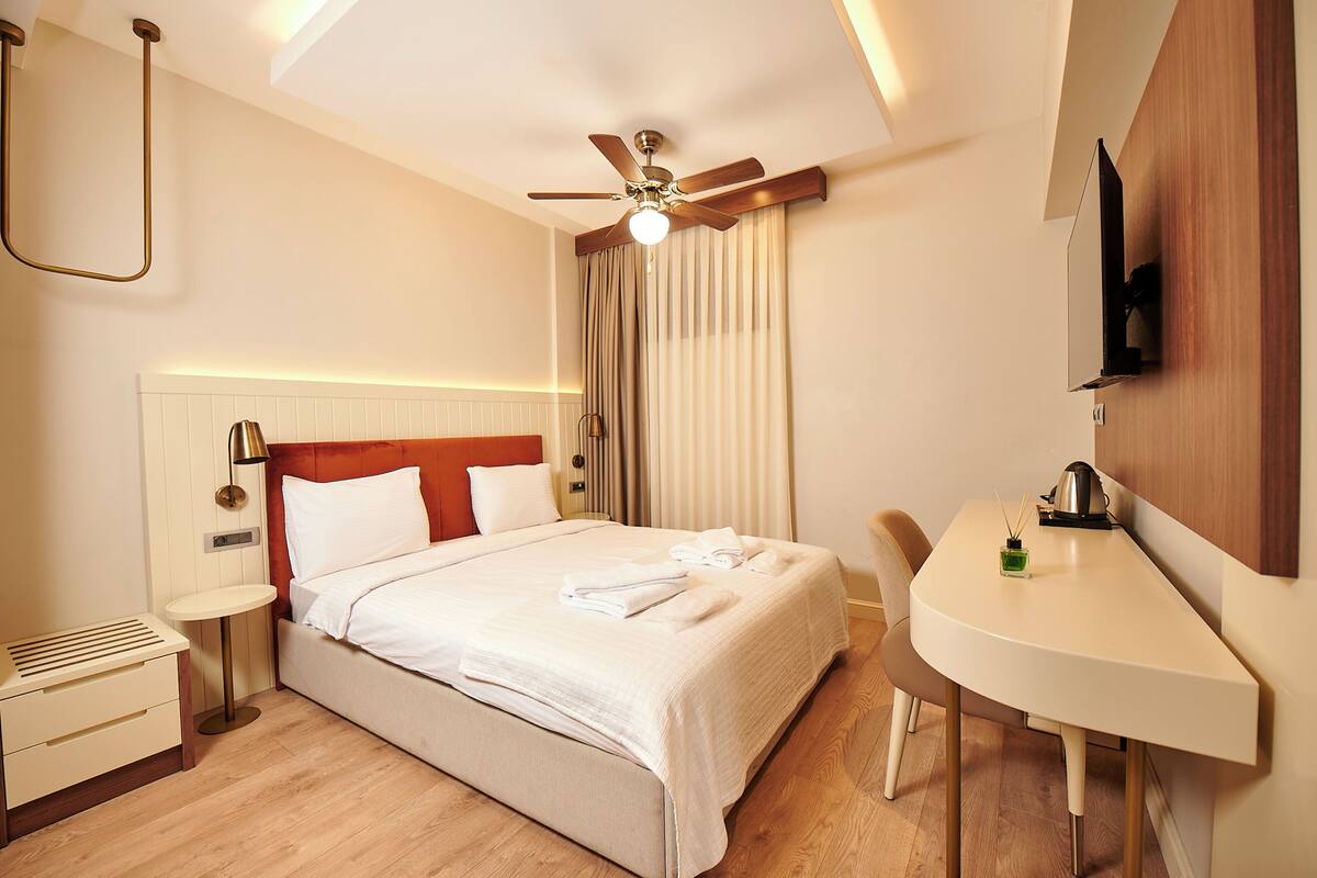 A hotel room with a bed, bedside table, desk, chair, and a ceiling fan.