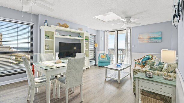A living room with a glass-top dining table, white wicker chairs, a teal armchair, a white sofa with a patterned throw, and a ceiling fan. - Recently Renovated Oceanfront Condo + Free Attraction Tickets! (North Myrtle Beach)